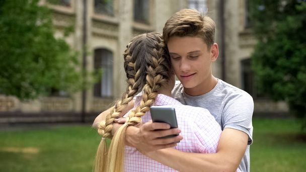 Teenager using cell phone while hugging girlfriend, absorbed by social networks - Valokuva, kuva