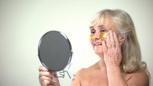 Joyful smiling lady applying eye patches looking in mirror, anti-aging therapy - Valokuva, kuva