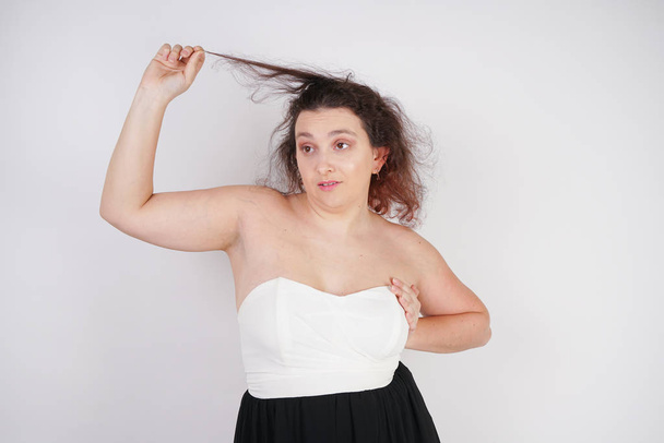 χαριτωμένο συναισθηματικό ενήλικος κορίτσι τραβάει ένα σκέλος των μαλλιών σε λευκό φόντο στο στούντιο - Φωτογραφία, εικόνα