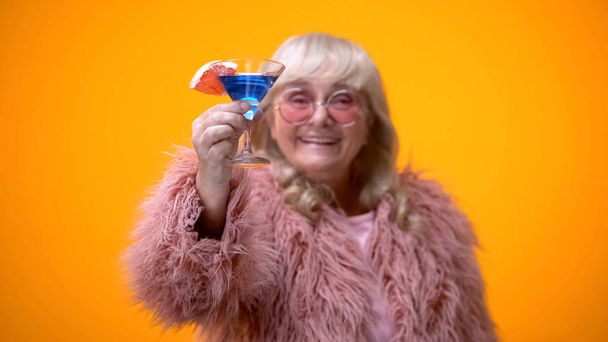 Cheerful aged woman in funny pink outfit raising toast, drinking blue cocktail - 写真・画像