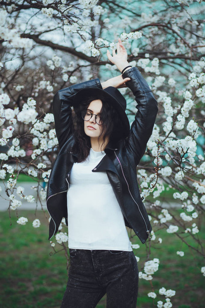 Outdoor spring portrait of fashion sensual young girl posing in amazing blooming garden. Wearing a hat and leather jacket, feminine look, elegant glamorous style. On the background of cherry blossoms - Foto, afbeelding