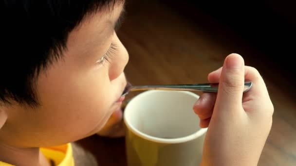 Ein kleiner Junge sitzt zu Hause am Holztisch am Fenster und trinkt eine Tasse heiße Schokolade. er hält die Tasse in der linken Hand und mischt heiße Schokolade mit Sahne mit einem Teelöffel in der rechten Hand. - Filmmaterial, Video