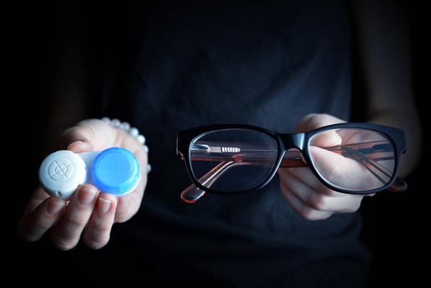 Hands holding contact lenses and glasses on a black background. - Photo, Image