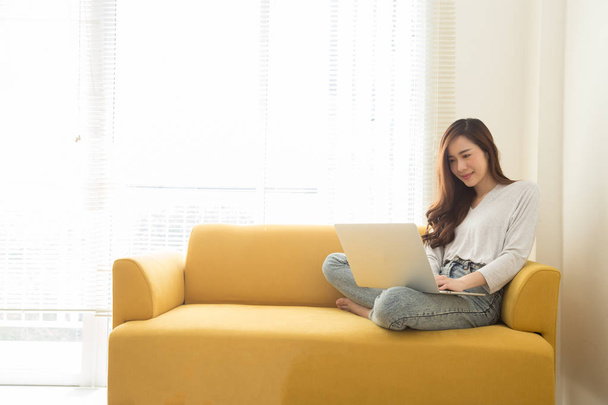 Young Asian woman sitting with laptop on yellow couch in home office. Concept of remote workplace and working at home - Fotografie, Obrázek