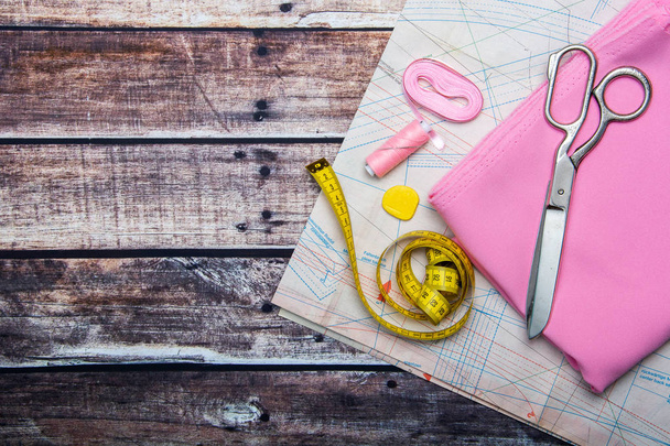 Tailoring. Naai benodigdheden en accessoires voor naaien en handwerk. Schaar, gele centimeter, patroon, spoel van draad en stof van roze kleur op een houten achtergrond. Platte lay, kopieer ruimte - Foto, afbeelding