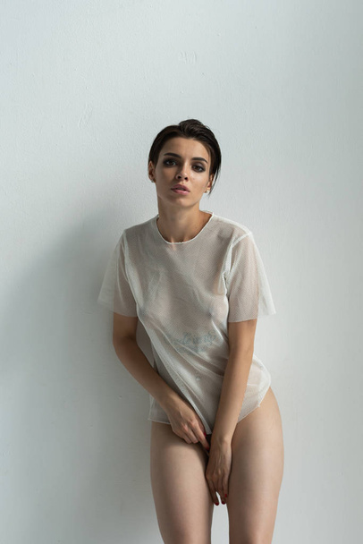 young beautiful girl with posing nude in studio standing in white t-shirt near white wall - Фото, зображення