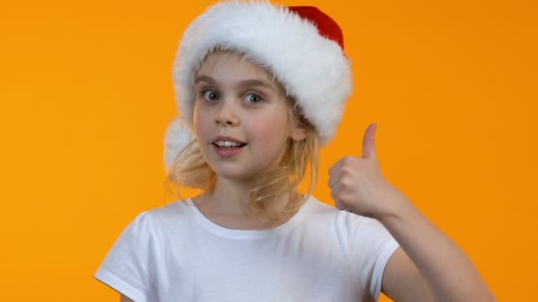 Little female kid in santa hat showing thumbs-up and winking on camera, holiday - Filmati, video