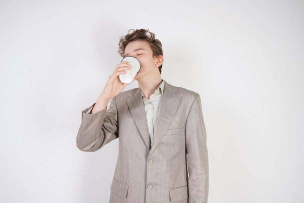 menino adolescente cansado em terno de negócios cinza com papel xícara de café na mão sobre fundo estúdio branco
 - Foto, Imagem