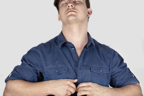 Young handsome caucasian man with dark curly hair and closed eyes unbuttons a blue jeans shirt. Hot sexy male. Grey background, studio, isolated, copy space. - Fotó, kép