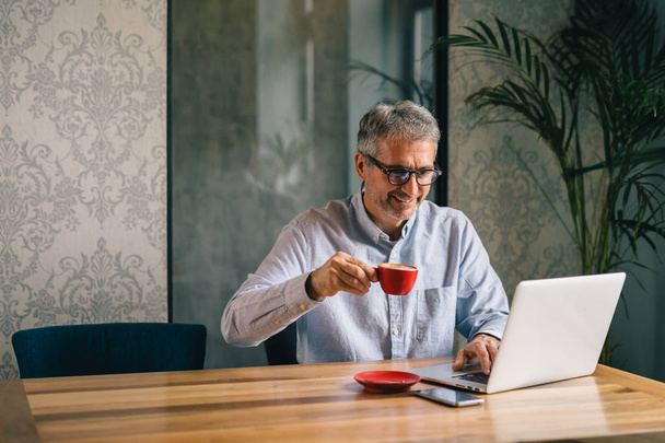 Smiling mature businessman using laptop and drinking coffee in office - Zdjęcie, obraz