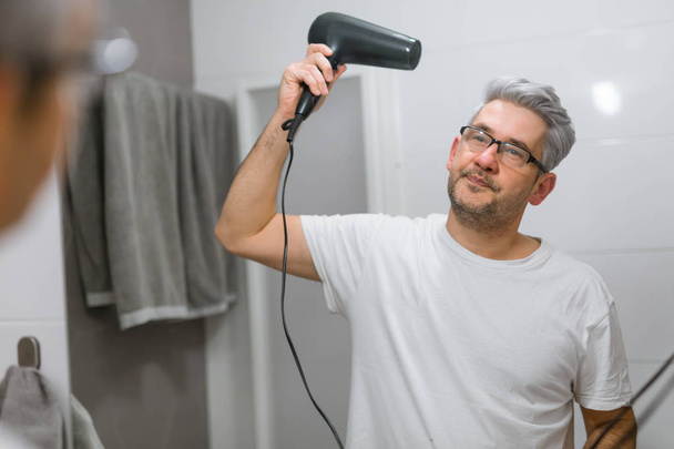 Gray haired man using hairdryer in bathroom - Foto, imagen
