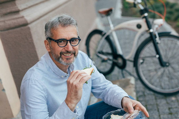 Mature man eating sandwiches outdoor in city - Foto, afbeelding