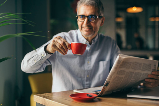Smiling businessman reading newspaper and drinking coffee in modern cafe bar. - Photo, Image