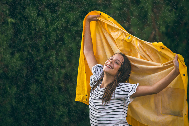 girl enjoying summer rainy days with yellow raincoat in rising hands - Photo, Image