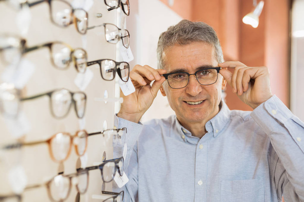 Mature man choosing the best eyeglasses in modern optic store - 写真・画像