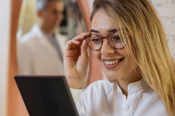 Close up of woman choosing glasses in optics store with optician on background - Photo, Image