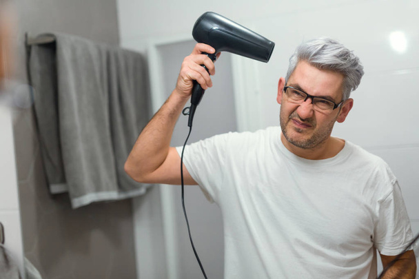 Gray haired man using hairdryer in bathroom - 写真・画像