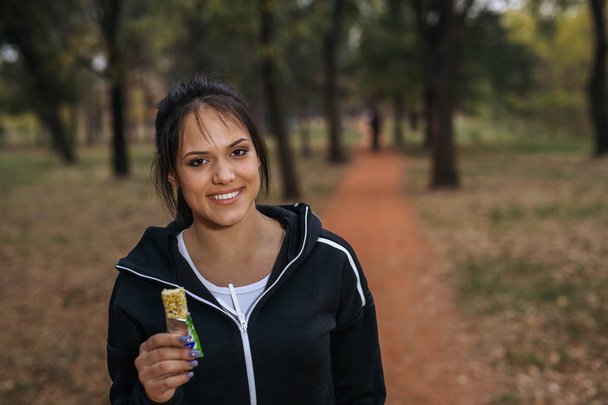 Porträt einer jungen Frau, die beim Joggen im Park Müsliriegel isst - Foto, Bild