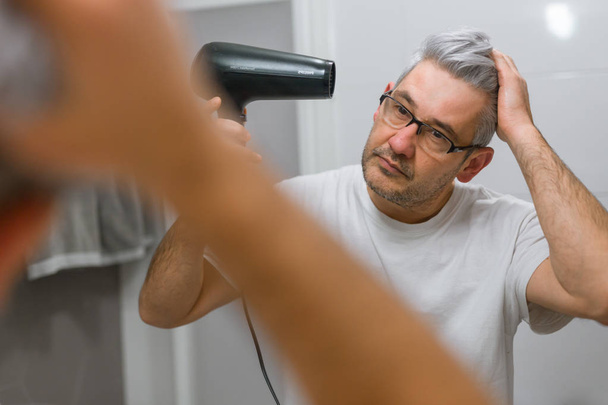Gray haired man using hairdryer in bathroom - Фото, зображення