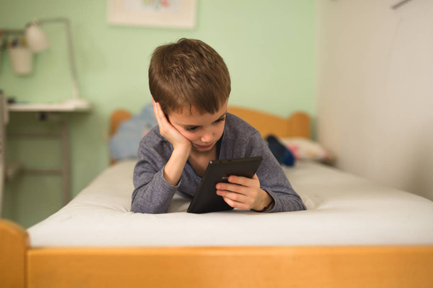 boy laying on bed in room and using tablet - Valokuva, kuva