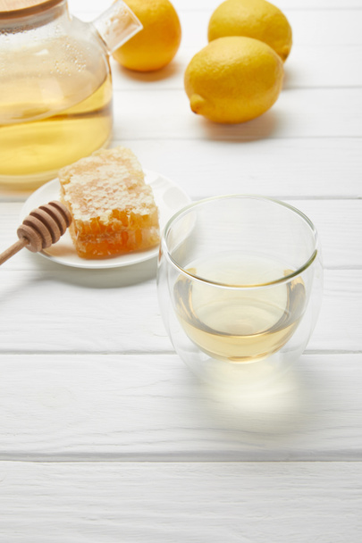 průsvitné sklo a čajník se zeleným čajem, citronem a voštovým pláčem na bílém dřevěném stole - Fotografie, Obrázek
