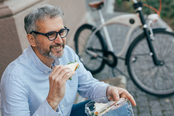 Mature man eating sandwiches outdoor in city - Fotoğraf, Görsel