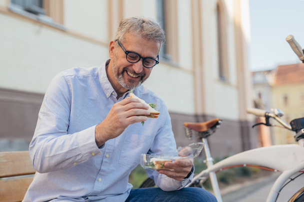 Mature man eating sandwiches outdoor in city - Foto, Imagem