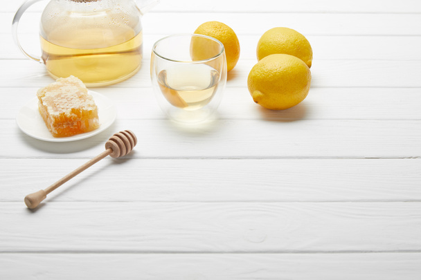 Průhledný čajový hrnec a sklo se zeleným čajem, citronem, voštovým a dřevěným medovým sušáčem na bílém stole - Fotografie, Obrázek