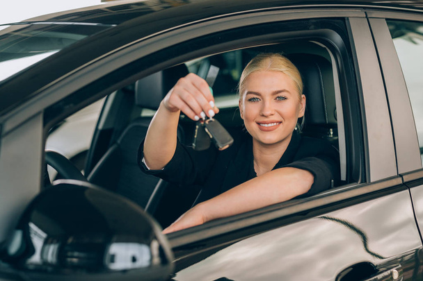 woman sitting in new car and holding keys in car dealership showroom - Foto, Imagen