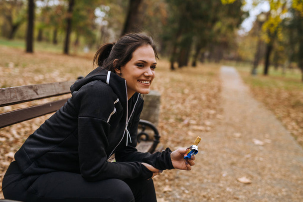 Woman jogger sitting in park and eating granola bar - 写真・画像