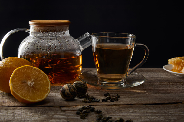 transparent teapot and cup with traditional blooming tea on wooden table with lemons isolated on black - Photo, image