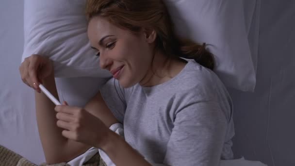Happy woman lying in bed, looking at pregnancy test with positive result - Séquence, vidéo