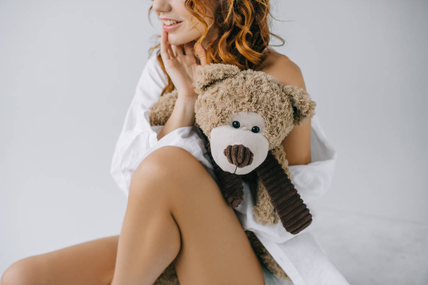 cropped view of cheerful curly young woman holding teddy bear while sitting on white  - Valokuva, kuva