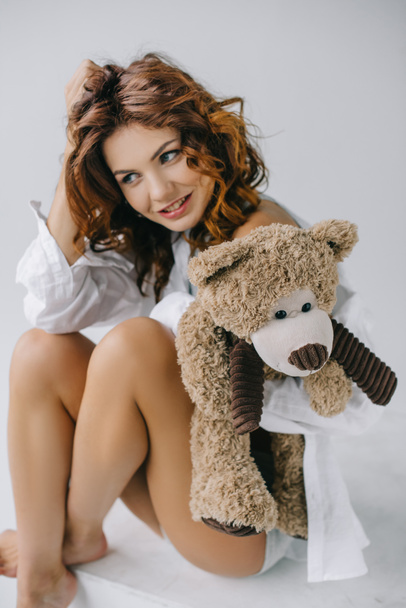 happy curly young woman holding teddy bear while touching hair on white  - 写真・画像