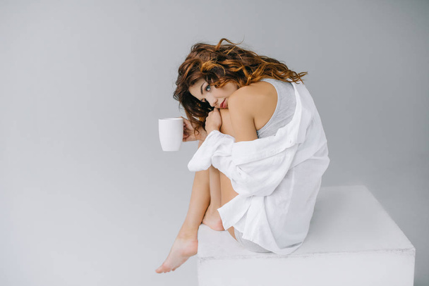 mujer joven soñadora y atractiva sosteniendo la taza con café en gris
  - Foto, Imagen