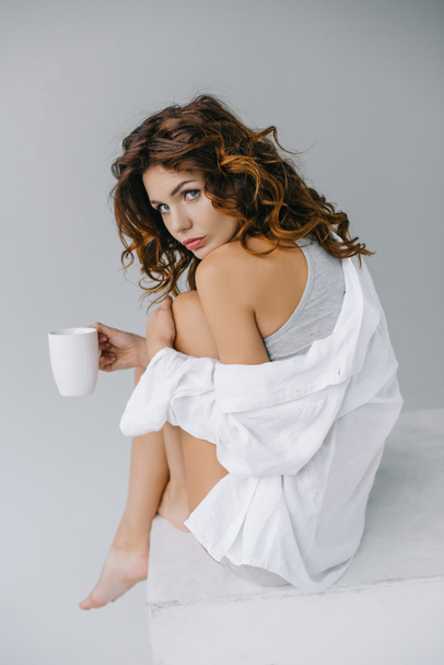 attractive young woman holding cup with coffee and looking at camera on grey  - Fotó, kép