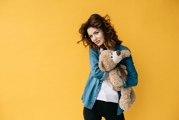 attractive redhead girl hugging teddy bear and looking at camera on orange  - Foto, immagini