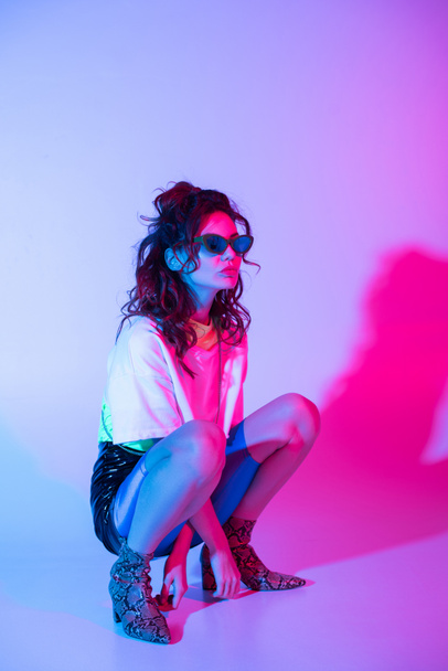 attractive and stylish girl in sunglasses sitting on purple with illumination - Foto, Imagem