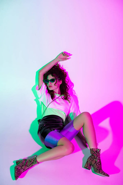 trendy beautiful young woman in sunglasses sitting on pink with illumination  - Fotografie, Obrázek