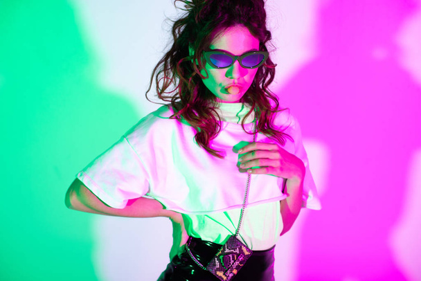 attractive girl in sunglasses blowing bubble gum on pink with illumination   - Фото, изображение