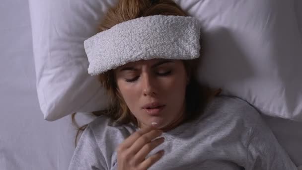 Woman lying in bed with towel on forehead, taking pills, cold treatment, illness - Materiał filmowy, wideo