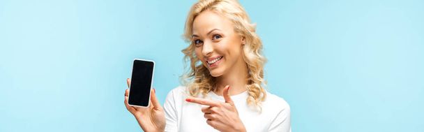 panoraama laukaus iloinen blondi nainen osoittaa sormella älypuhelin tyhjä näyttö eristetty sininen
 - Valokuva, kuva