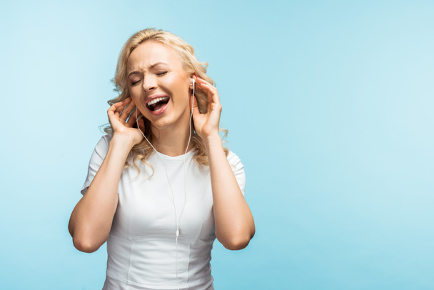 fröhliche Frau mit geschlossenen Augen, die mit Kopfhörern Musik hört und auf blau singt  - Foto, Bild