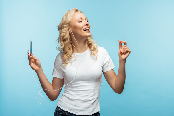 mujer feliz con los ojos cerrados escuchando música en los auriculares mientras sostiene el teléfono inteligente en azul
  - Foto, Imagen