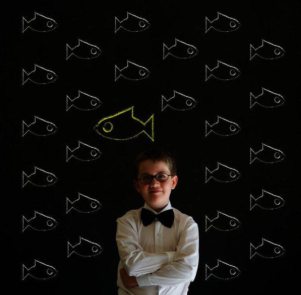 Thinking boy dressed up as business man with independent thinking chalk fish swimming against the flow on blackboard background - Fotoğraf, Görsel