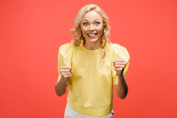 happy blonde woman looking at camera and celebrating success on red  - Foto, afbeelding