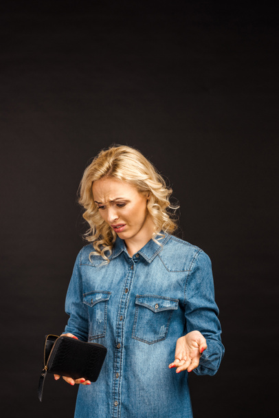 zdenerwowany blond kobieta patrząc na pusty portfel i wskazując izolowane na czarnym  - Zdjęcie, obraz