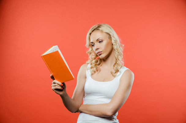attractive curly blonde woman reading book while standing on red  - Foto, Imagem