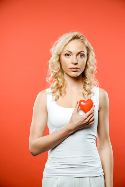 attractive blonde woman holding heart while looking at camera on red  - Foto, Bild