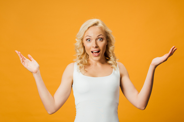 surprised blonde woman gesturing and looking at camera isolated on orange  - Фото, зображення